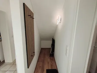 Ein heller Flur mit schrägem Dach und Holzverkleidung. Der Boden ist mit Holzplatten ausgelegt und es gibt eine Wandlampe.