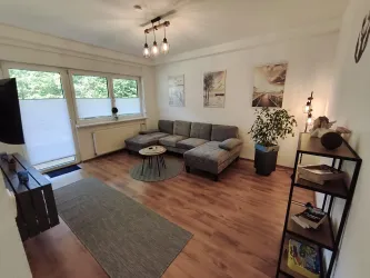 Ein modern eingerichtetes Wohnzimmer mit einer grauen Couch und einem kleinen Tisch. Große Fenster bringen viel Licht und eine Pflanze sorgt für einen frischen Akzent.