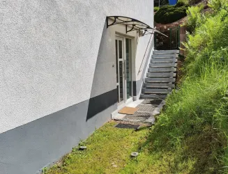 Ein Eingang zu einem Gebäude mit einer Treppe und einem kleinen Vorgarten. Die Wand ist hell gestrichen und das Gelände ist grün und gepflegt.
