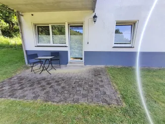 Eine kleine Terrasse mit einem Tisch und zwei Stühlen vor einem Haus. Der Garten ist grün und gut gepflegt.