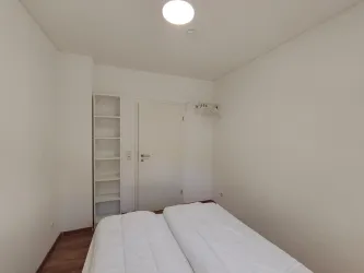 Ein schlichtes Schlafzimmer mit zwei Einzelbetten und hellem Holzfußboden. An der Wand befindet sich ein offener Schrank und eine Tür zu einem weiteren Raum.