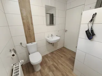 Ein modernes Badezimmer mit weißen Fliesen und einem Holzpaneel. Es enthält eine Toilette, ein Waschbecken und einen großen Spiegel.