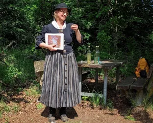 Eine Frau in traditioneller Kleidung steht in einer grünen Umgebung und hält ein Buch in der Hand. Auf einem Tisch neben ihr stehen zwei Flaschen und weitere Gegenstände.