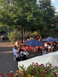 Eine belebte Außenbereich mit mehreren Tischen und Sonnenschirmen. Menschen genießen die Zeit im Freien unter einem großen Baum.