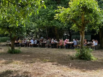 Eine Gruppe von Menschen sitzt an langen Tischen im Freien unter Bäumen. Die Atmosphäre ist entspannt und freundlich.
