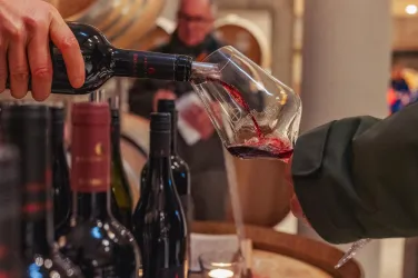 Ein Weinglas wird mit roten Wein gefüllt. Im Hintergrund sind unscharfe Personen und Flaschen zu sehen.