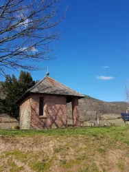 Eine kleine, gemauerte Kapelle steht auf einem grasbewachsenen Hügel. Im Hintergrund sind sanfte Hügel und ein klarer blauer Himmel zu sehen.