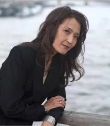 Eine Frau in einem schwarzen Blazer steht am Wasser. Ihr Blick ist nachdenklich und sie hat lange, lockige Haare.