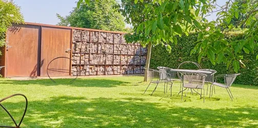 Ein schöner Garten mit grünem Rasen und einem Tisch mit Stühlen. Im Hintergrund steht eine Holzschuppen und es sind Bäume zu sehen.