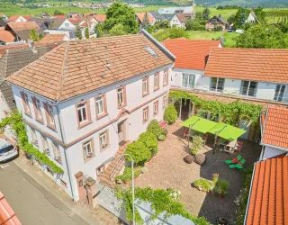 Eine charmante Vorderansicht eines Hauses mit einem begrünten Innenhof. Im Hof befinden sich Sitzgelegenheiten und Sonnenschirme, umgeben von Bäumen und Pflanzen.