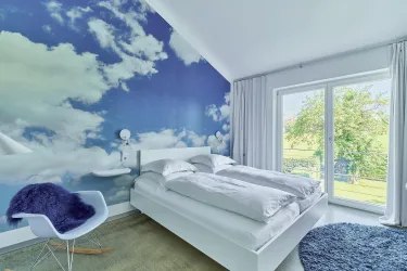 Ein modernes Schlafzimmer mit einem Himmel-Motiv an der Wand. Große Fenster sorgen für viel Licht und einen schönen Ausblick ins Grüne.