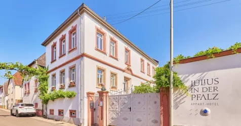 Ein charmantes Apartment-Hotel mit einem hellen Gebäude und grünen Pflanzen. Das Wetter ist sonnig und der Himmel ist blau.