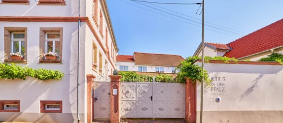 Ein charmantes Gebäude mit einer weißen Fassade und bunten Blumen an den Fenstern. Das Tor zum Grundstück ist schön verziert und umgeben von Pflanzen.