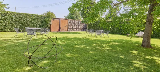 Eine grüne Wiese mit mehreren Tisch- und Stuhl-Gruppen im Freien. Im Hintergrund steht eine Holzstruktur und es gibt eine künstlerische Skulptur aus Metall.