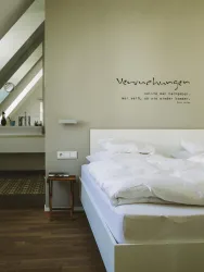 Ein modernes Schlafzimmer mit einem großen Bett und weißen Bettdecken. An der Wand steht ein Zitat, und im Hintergrund ist ein Waschbecken zu sehen.