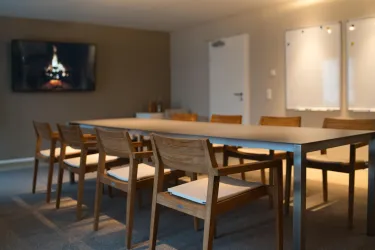 Ein moderner Besprechungsraum mit einem langen Tisch und Holzstühlen. An der Wand ist ein Fernseher, der ein Kaminfeuer zeigt.
