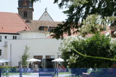 Ein modernes Gebäude mit einer weißen Fassade, umgeben von Bäumen und einem schönen Garten. Im Hintergrund ist ein Turm einer Kirche sichtbar.