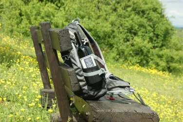 Ein Rucksack mit einer Wasserflasche steht auf einer Bank inmitten einer blühenden Wiese. Der Hintergrund zeigt frisches Grün und eine friedliche Landschaft.