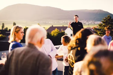 Eine Feier auf einer Dachterrasse mit Menschen, die sich versammeln und miteinander sprechen. Im Hintergrund ist eine malerische Landschaft und ein hügeliges Terrain zu sehen.