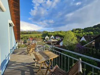 Ein Balkon mit Holzmöbeln bietet einen Blick auf eine malerische Landschaft. Im Hintergrund sind grüne Hügel und kleine Häuser sichtbar.