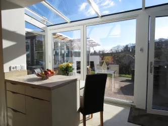 Eine helle Küche mit großen Fenstern und Blick auf eine grüne Landschaft. Ein Tisch ist dekoriert mit Obst und einer Blumenvase.