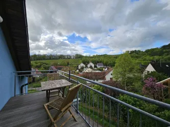 Ein Balkon mit einem Holztisch und einem Stuhl bietet einen Blick auf grüne Weinberge und Häuser. Der Himmel ist bewölkt, was eine ruhige Atmosphäre schafft.
