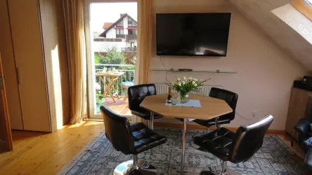 Ein heller Raum mit einem Tisch und schwarzen Stühlen. Im Hintergrund sind ein Fernseher und ein Balkon mit Aussicht auf einen Garten.