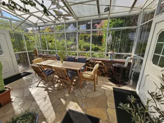 Ein heller Wintergarten mit einem großen Tisch und Holzstühlen. Umgeben von Pflanzen und einer schönen Aussicht in den Garten.