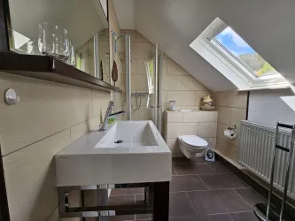 Ein modernes Badezimmer mit schrägen Wänden und einem Dachfenster. Es ist mit einem Waschbecken, einer Toilette und einem Spiegel ausgestattet.