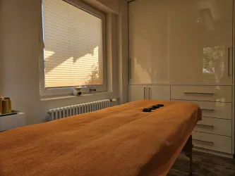 Ein ruhiger Behandlungsraum mit einem orangefarbenen Massagetuch und einem großen Fenster. Einige Steine liegen auf dem Tisch, was auf eine Massagebehandlung hindeutet.
