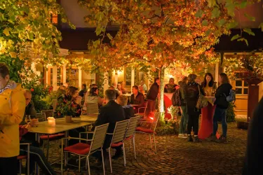 Eine lebhafte Terrasse mit vielen Gästen unter leuchtenden Bäumen. Es ist Abend, und die Atmosphäre ist gemütlich und einladend.