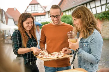 Drei Personen stehen unter einem Vordach und genießen gemeinsam Essen. Sie lächeln und haben Getränke in der Hand.