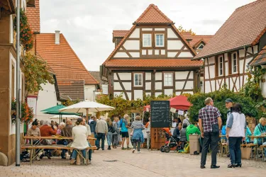Eine lebhafte Straßenszene mit Menschen, die an Tischen sitzen und sich unterhalten. Im Hintergrund sind traditionelle Fachwerkhäuser zu sehen.