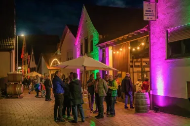 Eine lebhafte Veranstaltung bei Nacht mit Gäste, die sich vor einem beleuchteten Gebäude versammeln. Die Atmosphäre ist festlich mit bunten Lichtern und Schirmen.