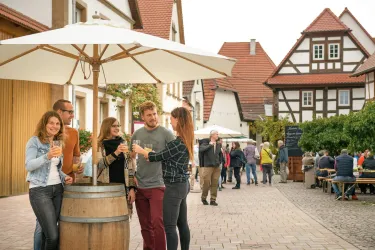 Ein belebter Marktplatz mit Menschen, die sich unter einem Sonnenschirm versammeln. Im Hintergrund sind traditionelle Häuser und Stände.