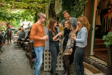 Eine Gruppe von fünf Freunden steht lächelnd in einer gemütlichen Gasse und genießt Drinks. Im Hintergrund sind weitere Gäste an Tischen zu sehen, umgeben von Pflanzen und einem einladenden Ambiente.