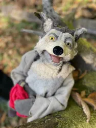 Eine Handpuppe eines Wolfes mit grauem Fell und einem freundlichen Gesichtsausdruck. Die Puppe hält ein rotes Tuch in der Hand und ist auf einem Baumstamm platziert.