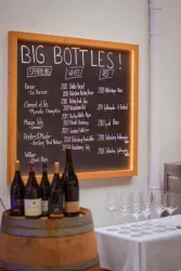 Eine Tafel mit der Aufschrift "BIG BOTTLES!" zeigt eine Liste von Weinen in verschiedenen Kategorien. Im Vordergrund stehen einige Flaschen Wein und Weingläser auf einem Tisch.