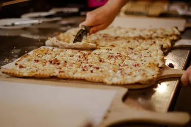 Ein frisch gebackenes Pizza, das in Stücke geschnitten wird. Im Hintergrund sind weitere Pizzastücke auf einer Holzplattform zu sehen.