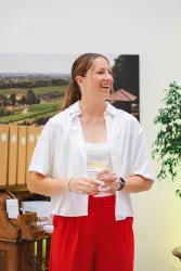 Eine junge Frau mit langen Haaren lächelt und hält ein Getränk in der Hand. Im Hintergrund sind Pflanzen und eine landschaftliche Szenerie sichtbar.