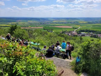 Eine Gruppe von Menschen sitzt an einer Aussichtsterrasse mit Blick auf grüne Felder und Hügel. Der Himmel ist blau mit einigen Wolken.