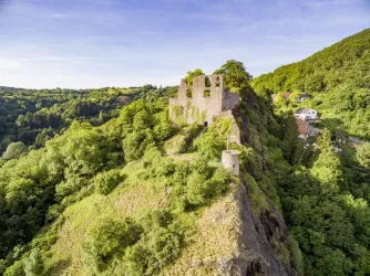 Eine malerische Ruine auf einem Hügel, umgeben von dichten Wäldern. Im Hintergrund sind grüne Landschaften und einige Wohnhäuser sichtbar.