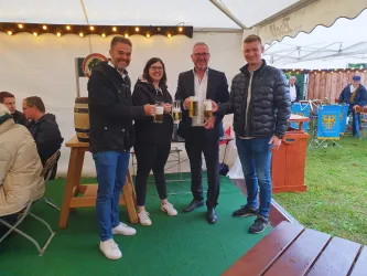 Eine Gruppe von vier Personen steht in einem Festzelt und hält Bierkrüge in den Händen. Im Hintergrund sind weitere Gäste und Festzeltmobiliar zu sehen.