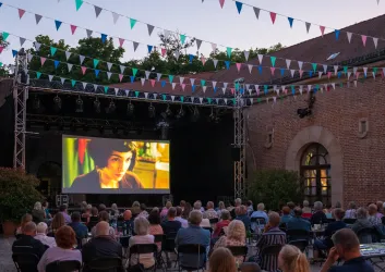 Ein Freiluftkino mit vielen Zuschauern, die auf eine große Leinwand schauen. Bunte Fahnen sind über den Platz gespannt, während die Dämmerung einsetzt.