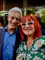 Ein lächelndes Paar steht vor einem Gebäude. Die Frau hat rote Haare und eine auffällige Brille, während der Mann einen blauen Hemd trägt.