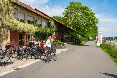 Ein gemütlicher Radweg neben einem Gebäude mit Pflanzen und einer großen Terrasse. Mehrere Fahrräder sind abgestellt, und der Himmel ist klar und blau.