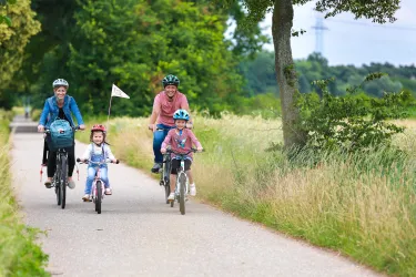 Eine Familie fährt gemeinsam mit dem Fahrrad auf einem schönen Weg. Es sind zwei Erwachsene und zwei Kinder, umgeben von grünen Bäumen und Wiesen.