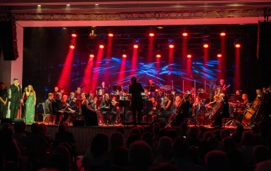 Ein Orchester spielt auf einer Bühne mit bunten Lichtstrahlen. Zwei Sänger stehen im Vordergrund und präsentieren eine beeindruckende Darbietung.