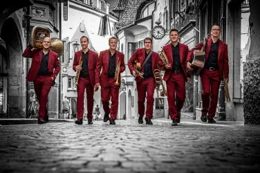 Eine Gruppe von sechs Musikern in roten Anzügen geht fröhlich eine historische Straße entlang. Sie halten verschiedene Musikinstrumente in den Händen und haben einen energiegeladenen Ausdruck.