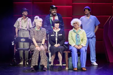 Eine Gruppe von sechs Personen in bunten Kostümen sitzt auf einer Bank auf der Bühne. Sie scheinen Teil einer Theateraufführung zu sein und schauen in die gleiche Richtung.